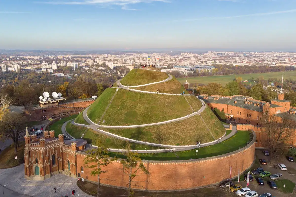Kopiec Kościuszki w Krakowie, zielona skarpa z krętą ścieżką, otoczona ceglanymi fortami. W tle rozciąga się panorama starego miasta.