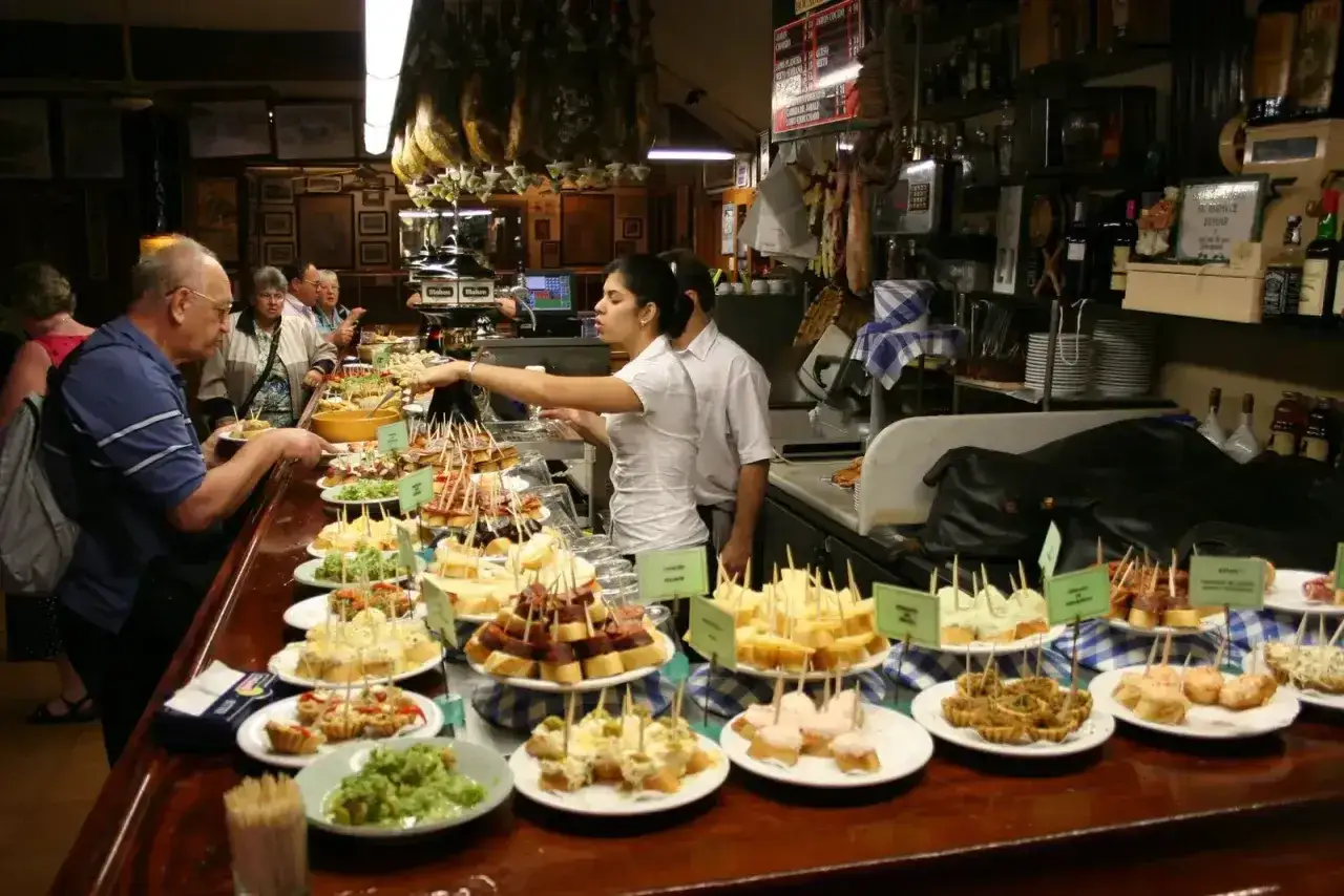 Tapas bar pełen ludzi wybierających przekąski. Na ladzie piętrzą się kolorowe pintxos, a za nią pracownicy obsługują gości.