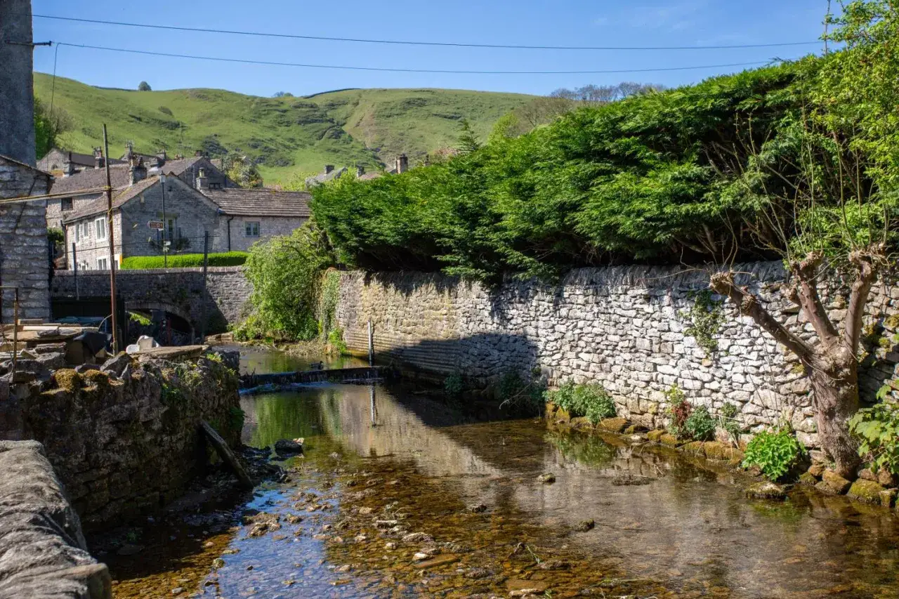 Kamienny mostek nad strumieniem w urokliwej wiosce w Cotswolds. Zielone wzgórza w tle.