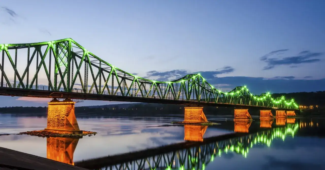 Zielony, oświetlony most nad rzeką, idealny na wypad na weekend. Jego odbicie w wodzie tworzy magiczny widok.