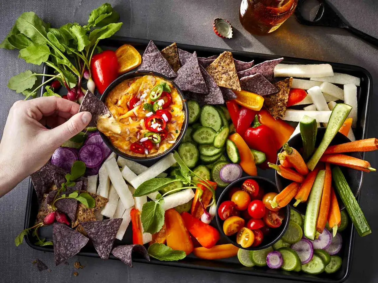 Szybka przekąska: ręka zanurza nachos w gorącym serowym dipie, otoczonym świeżymi warzywami i chrupiącymi chipsami. Idealne na imprezę!