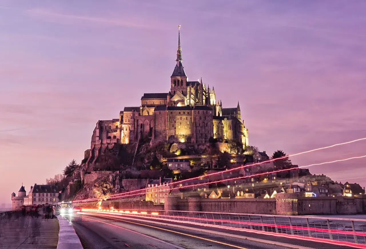 Mont Saint-Michel nocą, z rozświetloną architekturą i smugami świateł samochodów na drodze. Ciekawe ciekawostki o Francji!