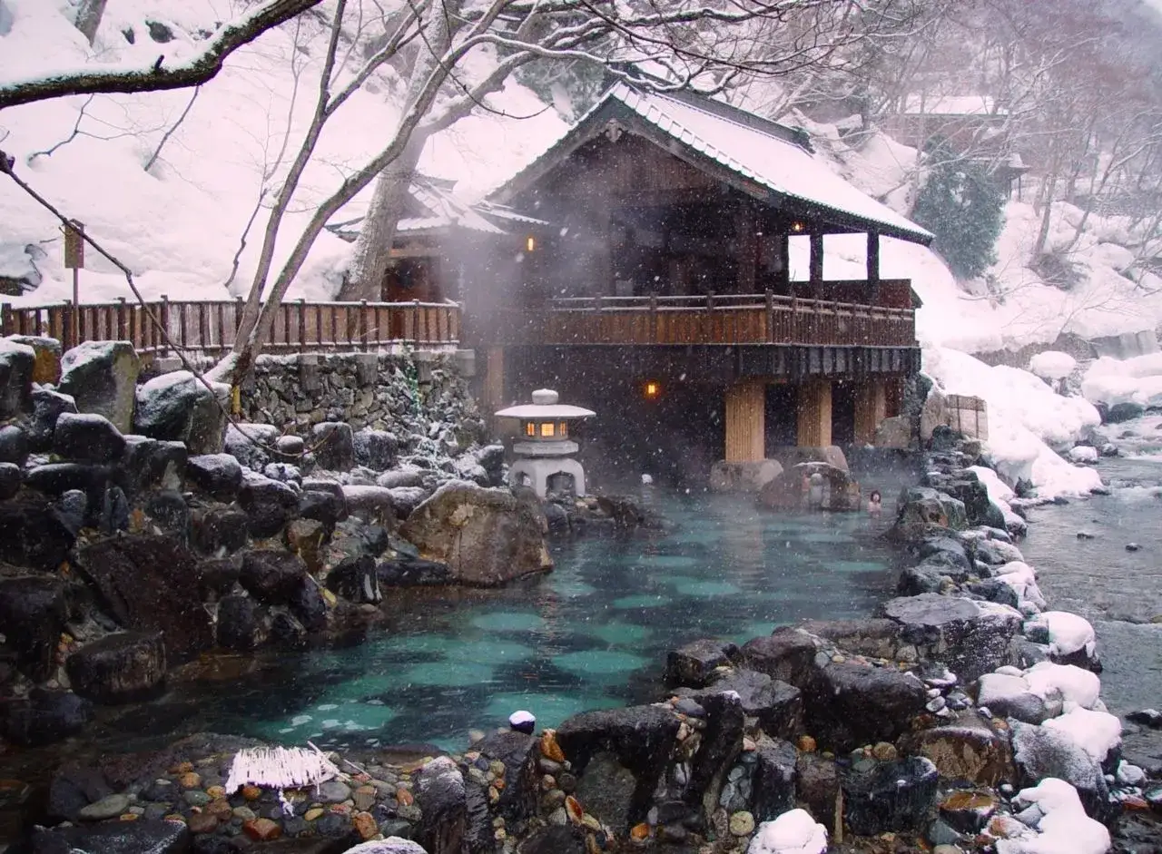 Zimowy onsen z parującą wodą, kamieniami i drewnianym budynkiem.