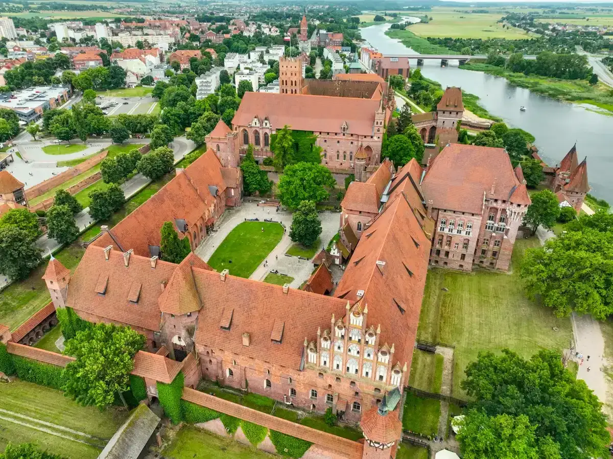 Ogromny zamek w Malborku, z czerwonymi dachami i zielonymi dziedzińcami, otoczony miastem i rzeką. Poznaj zamek w Malborku ciekawostki!