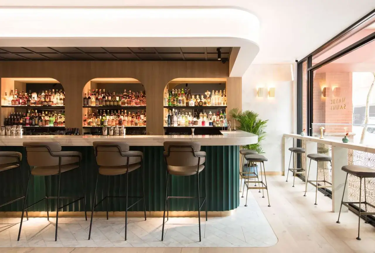 Elegancki bar z półkami pełnymi butelek, sugerującymi bogaty wybór popularnych drinków. Wygodne hokery zapraszają do degustacji.